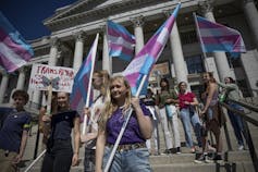 A group of people hold blue, pink and white striped flags and protest signs outside a white, Grecian-style building
