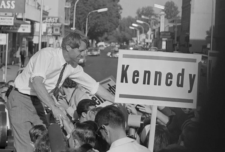 A man in shirtsleeves standing on a car, reaching out to shake people's hands.