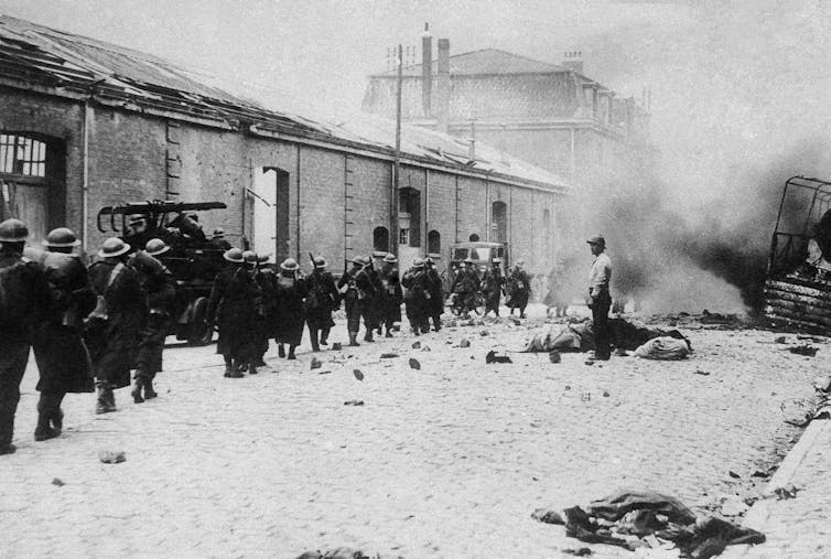 Troops marching along a street that has smoke rising at the end.