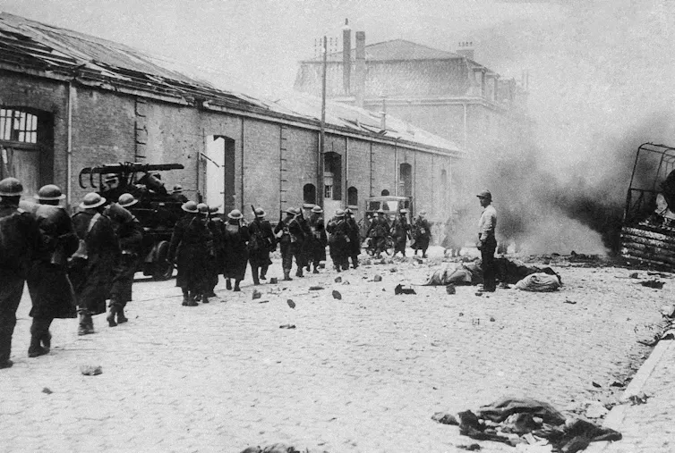 Troops marching along a street that has smoke rising at the end.