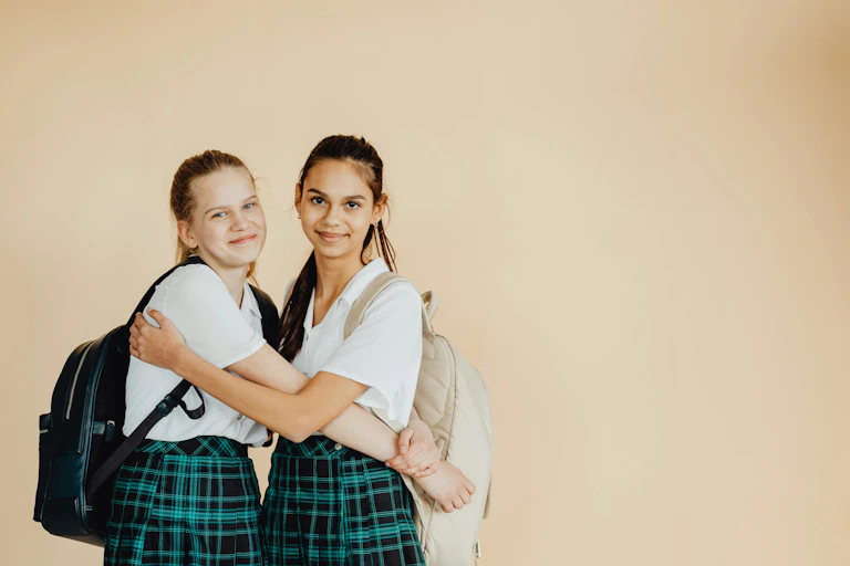 Two teenage girls in school uniforms and backpacks hug and smile.