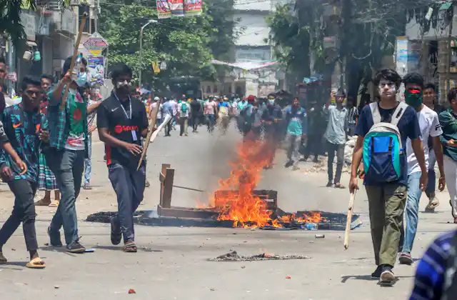 Protestors in Bangladesh walk down a street in front of a burning fire.