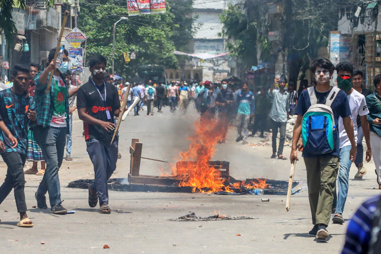 Protestors in Bangladesh walk down a street in front of a burning fire.