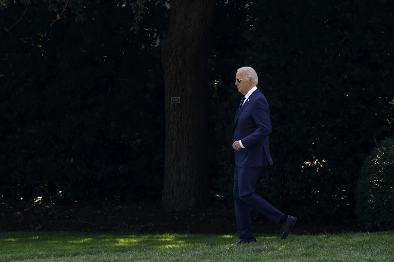 A man in a blue suit wearing aviator sunglasses walks in front of trees.