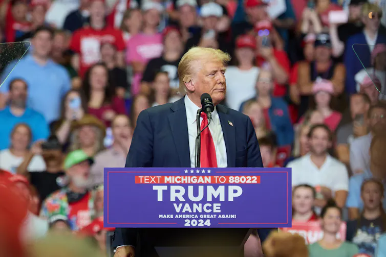 Donald Trump stands at a podium with Trump Pence and the number that donors need to call to give money. In the background are a crowd of supporters