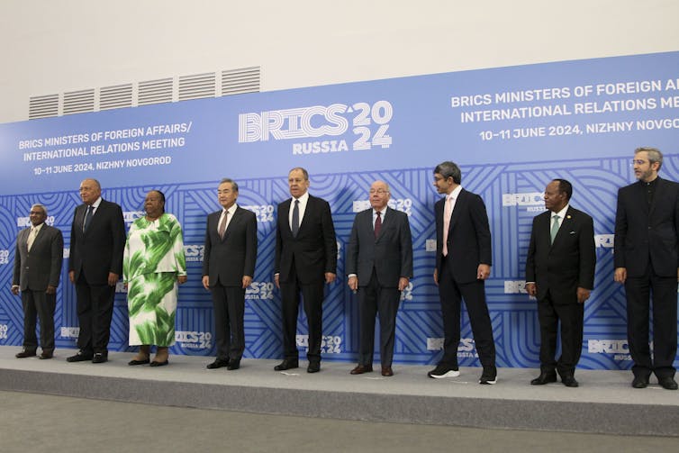 A group of people, all but one of them men, stand in a line on a stage posing for a photo.