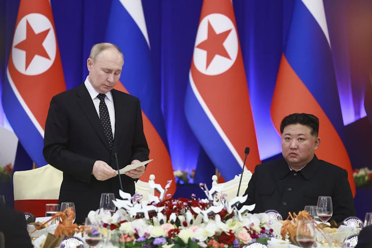 A balding man stands as he reads from notes as an Asian man dressed in black sits next to him at a dinner table.