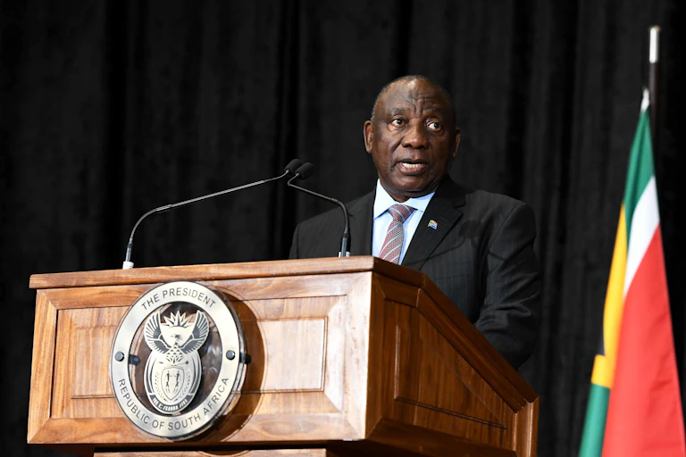 A man wearing a jacket and tie speaks at a podium bearing the South African coat of arms, next to the South African flag on his left.