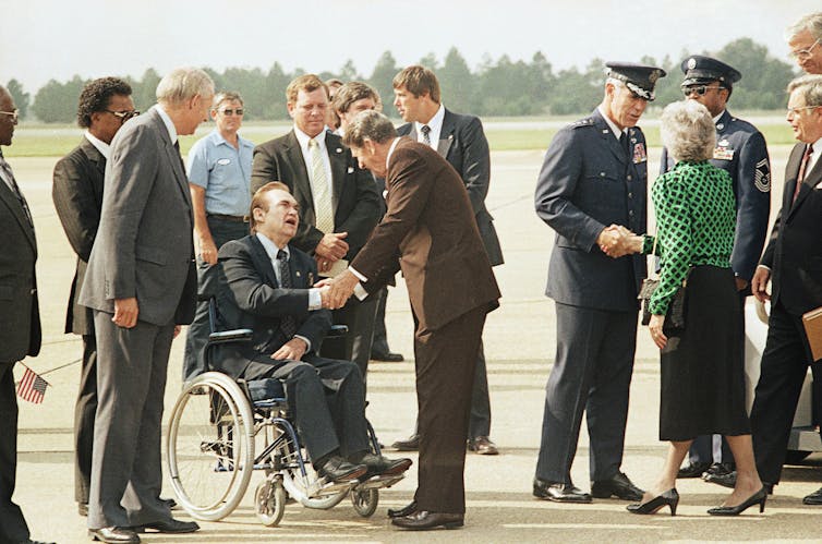 Reagan stands on a tarmac, bending to shake hands with Wallace seated in a wheelchair.