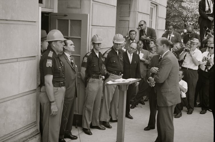 Wallace stands defiantly in a doorway surrounded by troopers and reporters.