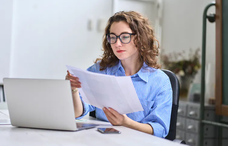 Une jeune femme lit une feuille de papier assise devant un ordinateur portable ouvert