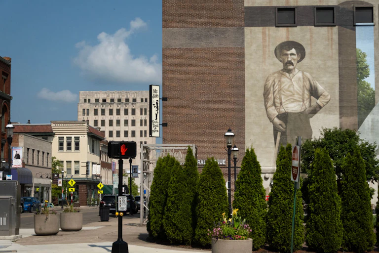 A sepia-toned mural of a man in suspenders and a white shirt, with a hat and a mustache, painted on the side of a multistory brick building.