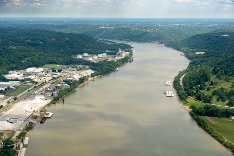 A wide, calm river between green, hilly areas, with some industrial buildings along one bank.