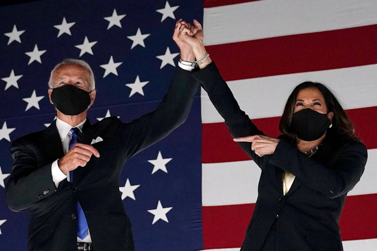 A man and a woman standing in front of a large American flag, wearing masks and holding raised hands.