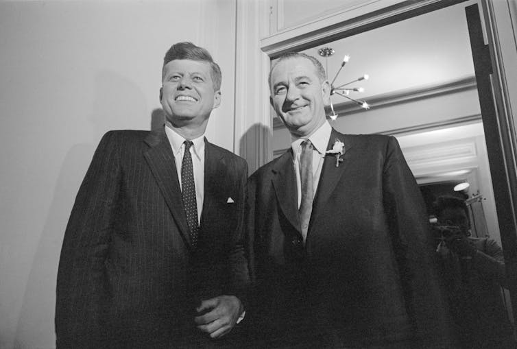 A dark-haired man stands next to a man with a receding hairline, both in suits and smiling.