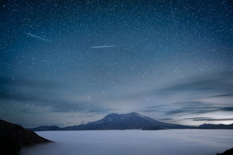 A misty mountain in the distance with a foggy lake in the foreground and several bright streaks in the sky.