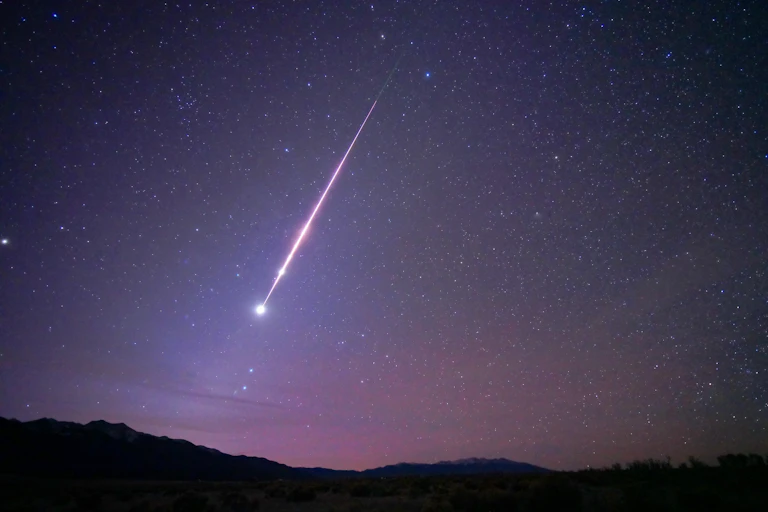 A bright fireball streaks across a purple-navy night sky towards a dark horizon.