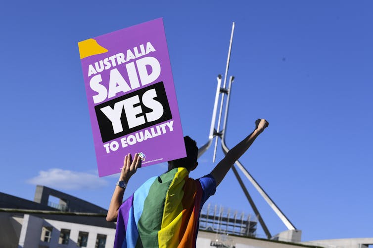 Man with LGBTIQ+ flag draped over shoulder 'Australia said yes to equality' placard, fist in air