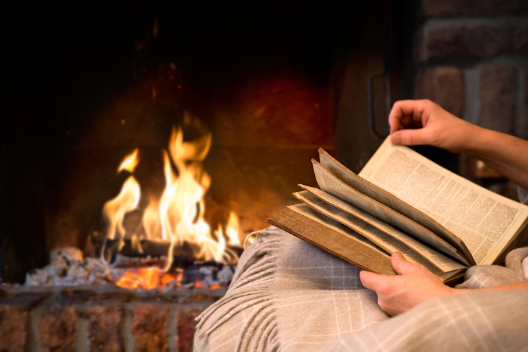 A person reads a book in front of an open fireplace