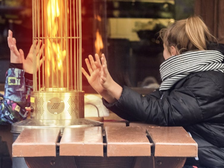 Two people warm their hands at an outdoor gas heater on a cafe table