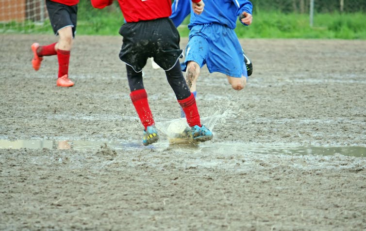Kids play football on a waterlogged field, which has been torn up and is now muddy.