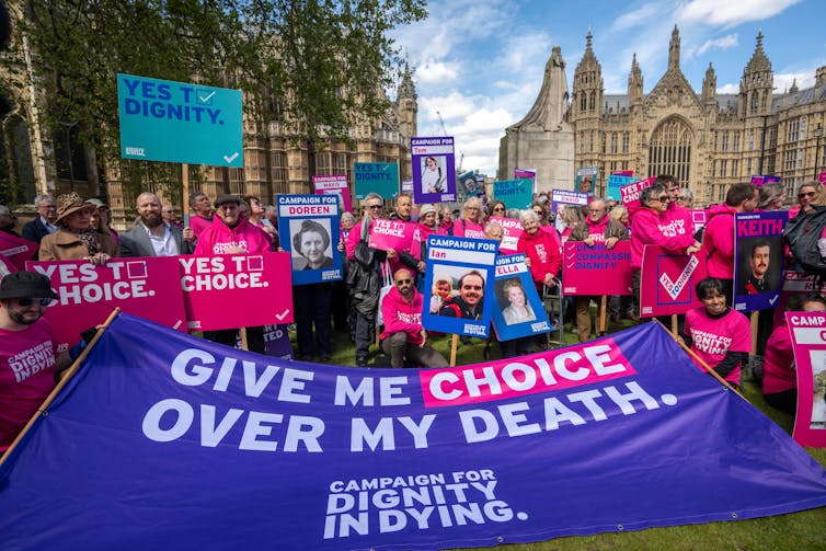 A group of campaigners standing near the Houses of Parliament holding a sign reading 'give me choice over my death'.