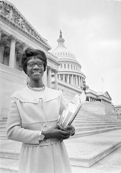 A black-and-white photo shows a Black woman wearing glasses and holding documents on Capitol Hill.