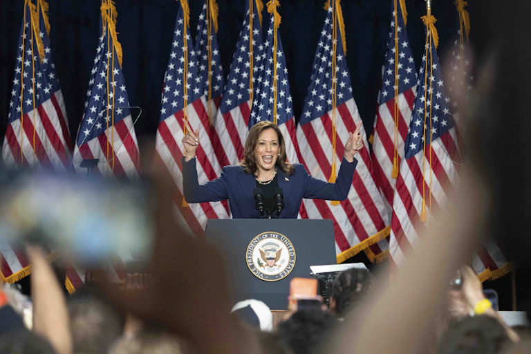 A smiling dark-haired woman raises her hands while speaking in front of a crowd, a row of American flags behind her.