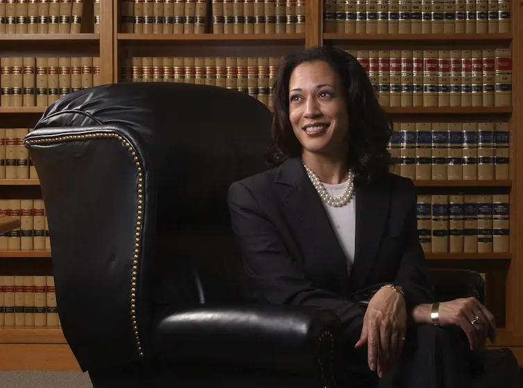 A dark-haired woman smiles sitting in a leather chair in front of large shelves of books.