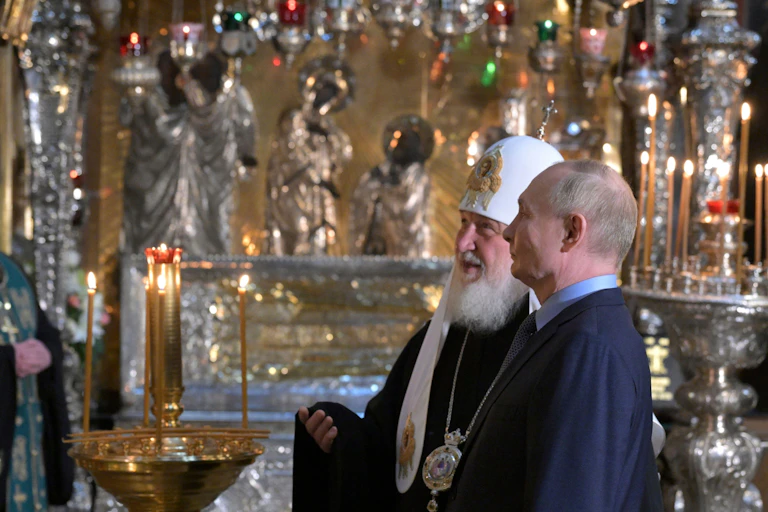 Russian president Vladimir Putin, right, accompanied by Russian Orthodox Patriarch Kirill visits to the Trinity Cathedral otuside Moscow. They are standing talking to each other.