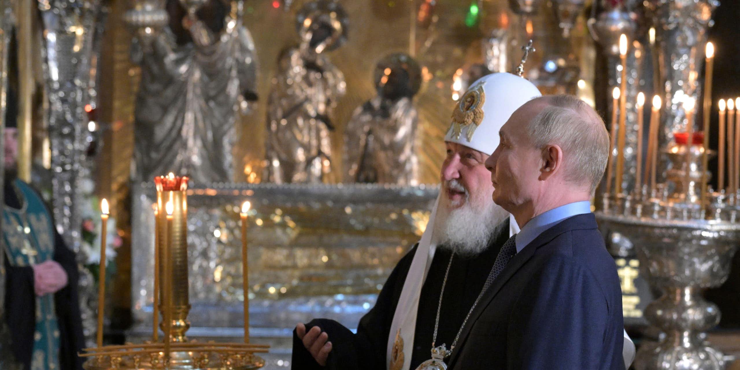 Russian president Vladimir Putin, right, accompanied by Russian Orthodox Patriarch Kirill visits to the Trinity Cathedral otuside Moscow. They are standing talking to each other.