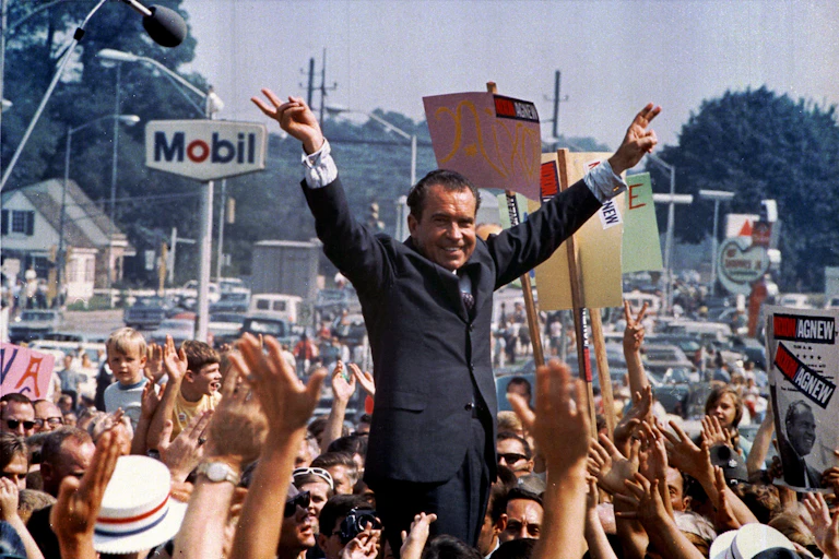 Richard Nixon raised above a crowd with both arms aloft.