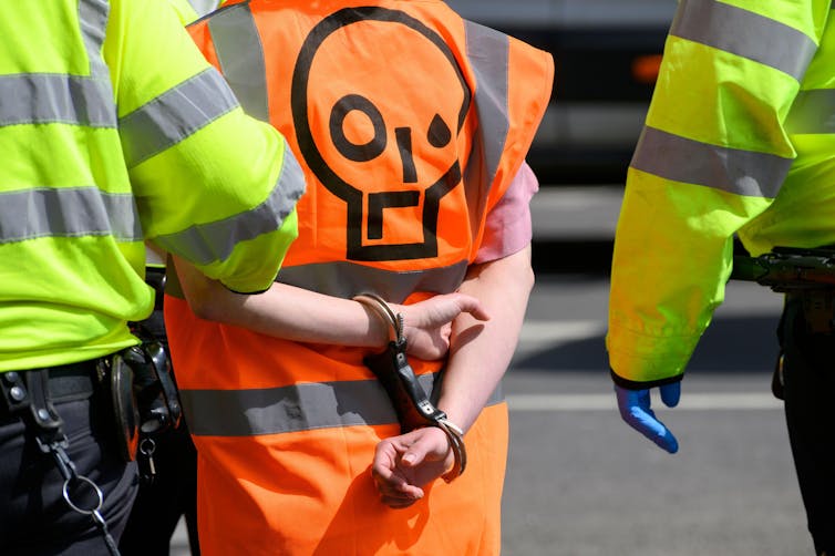 A person in orange hi-vis jacket is being led away in handcuffs by police.