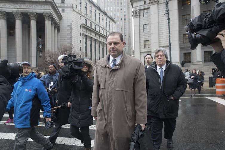 Joseph Percoco, wearing a trench coat, crosses a Manhattan street with court buildings in the background