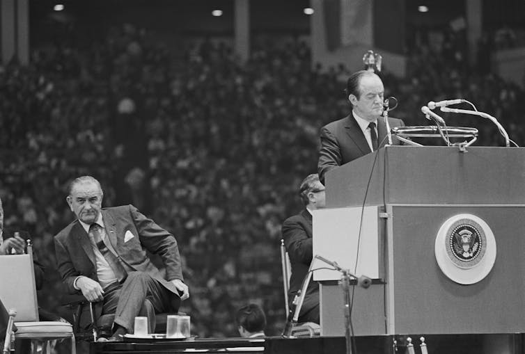 Two men at a large meeting, one in a chair and the other at a podium, speaking into a microphone.
