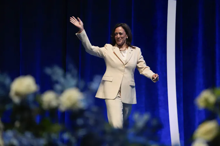 A dark-haired woman in a cream suit waves and smiles on a stage.