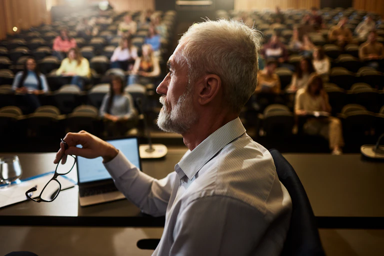 An older man holds eyeglasses while seated at a table inside a lecture hall.