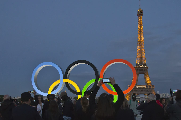 Os anéis olímpicos iluminados com a Torre Eiffel ao fundo. Pessoas em silhueta estão tirando fotos.