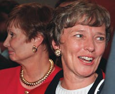 A woman with short blond hair smiles; another woman in a red jacket stands next to her, smiling and looking in the opposite direction.