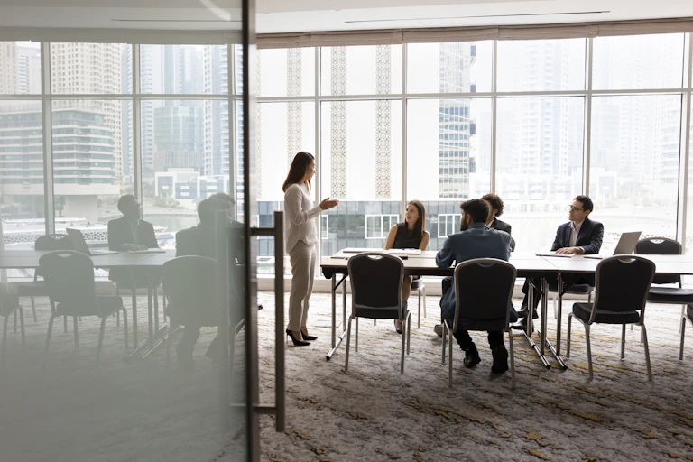Wide shot of a group of people sitting at a conference table, looking up at a woman who is speaking to them