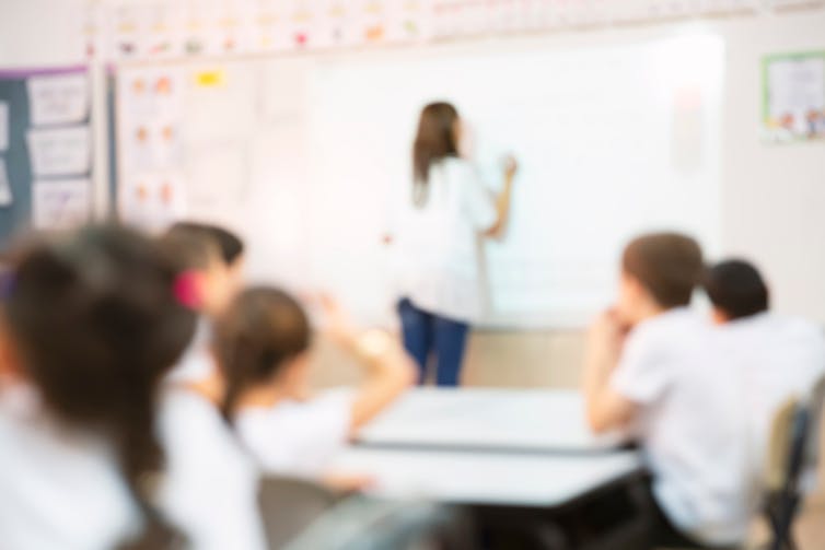 A blurred image of a teacher at a whiteboard with students in white uniform tops seated at desks.