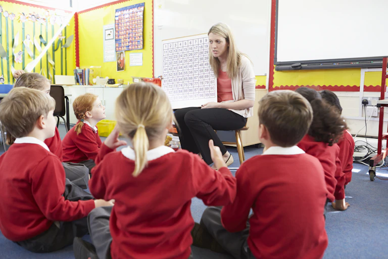 a teacher holds a board with numbers on it. Children sit in front of the teacher in a classroom.