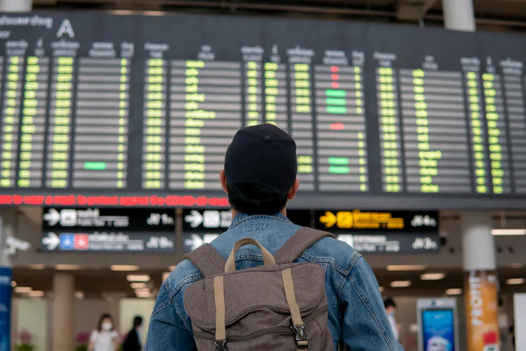 Back of young male traveler with backpack wearing a cap and looking at departure board in an airport terminal