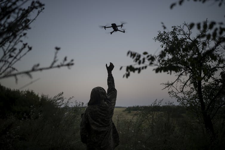 Photo at twilight of a person's silhouette reaching to grasp a descending quadcopter amid trees.