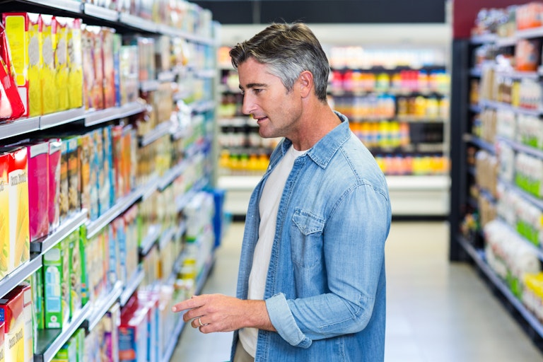 Middle aged man in supermarket aisle carrying shopping basket pointing at packaged goods on shelf