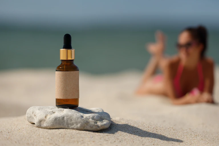 Une bouteille d’huile solaire sans étiquette posée sur une pierre, avec en arrière-plan une plage et une femme se faisant bronzer.