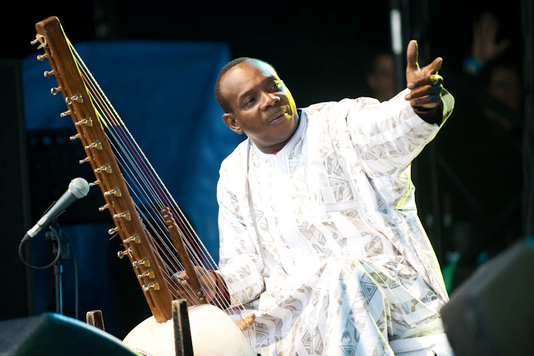 A man in an ornate white robe holds a hand up as he sits at a microphone with a large stringed musical instrument, apparently on a stage.