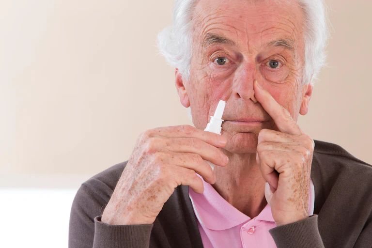 Elderly man holding a nasal spray bottle near his nose.
