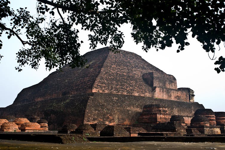 A large mound-like ancient stone structure, which appears blackened over the years, with a large tree near it.