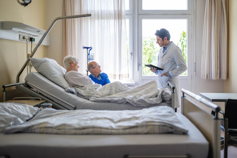In a hospital room, a doctor speaks with an older patient, with a male visitor sitting at bedside.